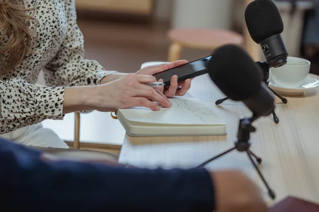 Crop anonymous with cellphone writing in planner while interviewing guest at table with mic and cup of coffee. Audience research for copywriting teams in England
